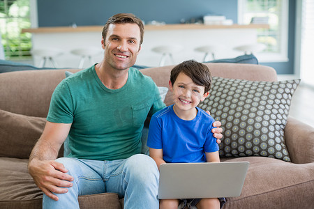 微笑的父亲和儿子坐在沙发上使用笔记本电脑的肖像