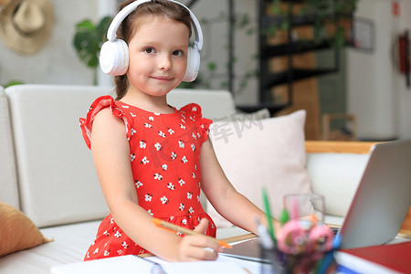 初等教育摄影照片_戴着耳机的微笑小女孩在家中使用笔记本电脑在线手写学习，戴着耳机的可爱快乐小孩子在电脑上上互联网网络课程或课程。