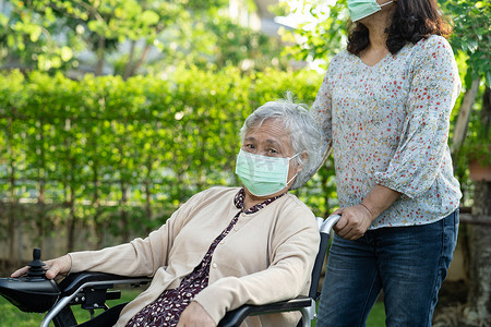 帮助坐电动轮椅和戴口罩的亚洲老年或老年老妇在公园里保护安全感染Covid-19冠状病毒。