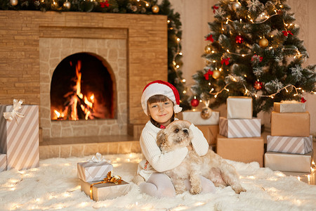 平安夜的小女孩和狗坐在白色柔软的地毯上，孩子看着相机拥抱她的宠物，戴着圣诞老人帽子的女孩坐在火炉和枞树附近。