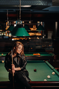 漂亮的桌面背景摄影照片_一个手里拿着球杆的女孩坐在台球俱乐部的桌子上。俄罗斯台球