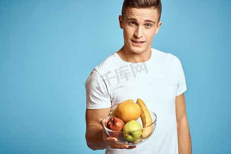 快乐的男人，手里拿着新鲜水果，双手蓝色背景白色T恤维生素香蕉橙子苹果