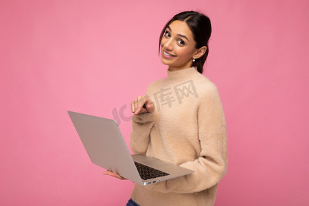 照片中，身穿米色毛衣的美丽黑发女子手持上网本电脑，看着相机，相机显示在粉色墙壁背景上隔离的笔记本电脑上