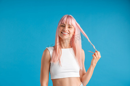 高兴的年轻女子，一头天然的粉色长发，手里拿着一缕头发，对着镜头微笑，在蓝色工作室背景下摆出孤立的姿势