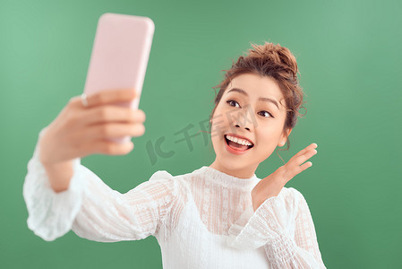 友好、外向的漂亮亚洲女性自拍或通过电话应用程序视频通话交谈，打个招呼，连接到直播，微笑着问候追随者，站立白色背景记录视频博客
