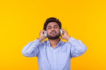 留着胡子的英俊轻松的印度男子正在黄色背景的耳机里听最喜欢的音乐。