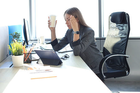 坐在工作地点，手里拿着纸杯的女商人看起来很疲惫的形象。