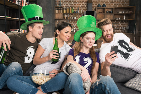 微笑的年轻朋友喝啤酒和使用智能手机，圣帕特里克节概念