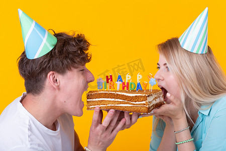 奇怪的年轻夫妇女孩和男孩戴着纸帽子的特写想要咬掉一块祝贺蛋糕。