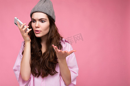 情感迷人的年轻黑发女性，穿着时尚的粉红色衬衫，戴着灰色帽子，在粉红色的背景下被隔离，手拿手机沟通和录制语音信息解释什么