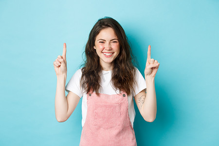 一张快乐微笑的卷发年轻女子的照片，她笑着用手指指着标志横幅，展示促销优惠，站在蓝色背景下