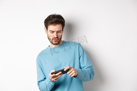 年轻人在智能手机上玩电子游戏，倾斜身体并点击手机屏幕，在白色背景中快乐地站着