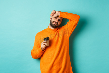 疲惫背景摄影照片_疲惫的成年男子下班休息，喝咖啡，擦去额头的汗水，看上去精疲力尽，站在绿松石背景下的橙色毛衣