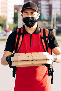 快递交付摄影照片_快递送货员的肖像，背着红色背包，拿着纸箱里的披萨，戴着防护面罩和手套。