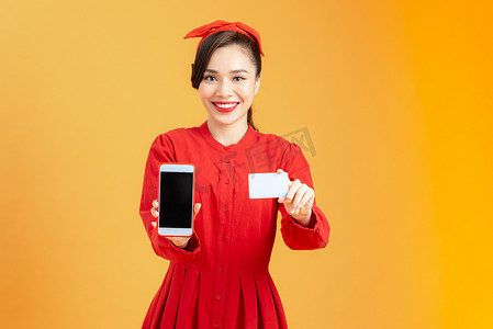 性格开朗的年轻亚洲女性站在橙色背景上，展示空白手机和信用卡。