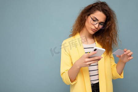 好看黄色摄影照片_身穿日常服装的年轻女性在背景中被隔离，手持电话和信用卡，通过信用卡支付网上购物费用，查看智能手机屏幕