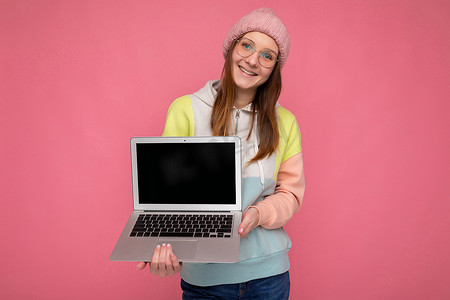 便携镜摄影照片_照片中，身穿帽子毛衣、戴着眼镜的漂亮年轻女士微笑着，手持电脑笔记本电脑，戴着白色耳机，看着粉红色背景中突显的相机