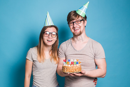 快乐的年轻夫妇迷人的男人和戴着纸帽子的可爱的女孩做出愚蠢的表情，手里拿着一个蛋糕，蛋糕上刻着生日，蓝色背景。