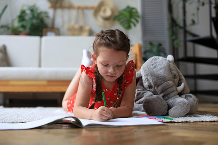 微笑快乐的女孩躺在温暖的地板上，身边有一只玩具大象，享受着创造性的活动，用铅笔为相册中的图片着色。