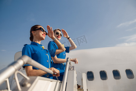两位身着蓝色制服、戴着墨镜、用手遮住眼睛、望向远方的优雅空姐的肖像，一起站在楼梯上