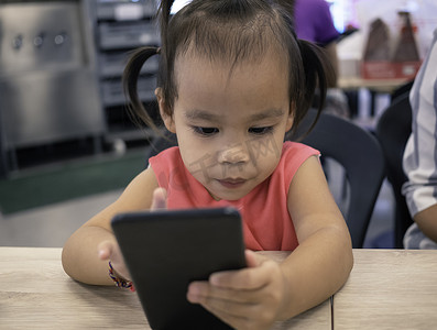 在购物中心的美食广场，亚洲小孩正在智能手机上愉快地看卡通片。