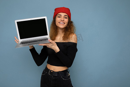 滑稽微笑、快乐、美丽的黑金发年轻女子的特写肖像，她拿着笔记本电脑，看着相机，身穿黑色作物上衣，红色和橙色的抹布隔离在浅蓝色墙壁背景上