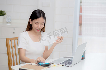 金融女性摄影照片_年轻的亚洲女性用家里桌上的计算器计算财务家庭，女孩检查账单并成功节省家庭开支、债务结清、税收和会计、商业理念。