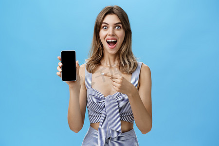 牙齿张开、兴奋、快乐的女人终于买了全新的智能手机手持设备，她指着手机屏幕，显示酷炫的应用程序，在蓝墙上快乐地微笑着