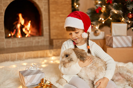 除夕夜，带着狗的女孩坐在壁炉旁的地板上，有北京犬作为礼物，非常高兴，穿着白色套头衫和红色圣诞老人帽子，孩子充满爱意地看着小狗。