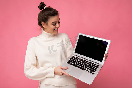 照片中，美丽迷人的微笑女子，黑发聚集，穿着白色毛衣，拿着电脑笔记本电脑，看着粉色墙壁背景上隔离的上网本
