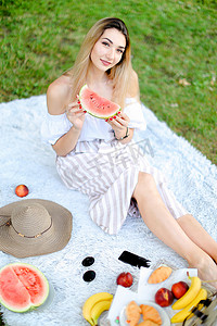 年轻漂亮的女孩坐在格子上，靠近水果和帽子，吃西瓜，背景是草。