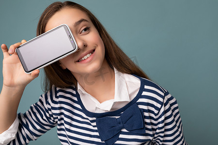 迷人的、积极微笑的黑发女孩，穿着条纹长袖，站在蓝色背景上，与世隔绝，复制空间拿着智能手机，手里拿着手机，屏幕显示空，用于模型指向 gadjet，看着旁边