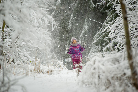 紫色衣服摄影照片_冬天，一个穿着紫色衣服的小女孩穿过白雪覆盖的森林