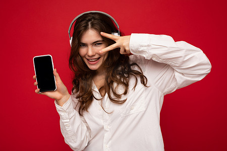 美丽快乐的微笑年轻女子穿着时尚休闲装，隔离在背景墙上，拿着手机，展示空显示屏，用于模型，戴着白色蓝牙耳机听音乐，开心地看着相机
