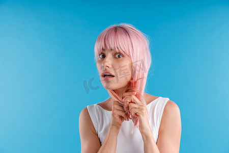 惊讶的女模特玩着粉红色的头发，在蓝色工作室背景下摆出孤立的姿势时触摸它的肖像