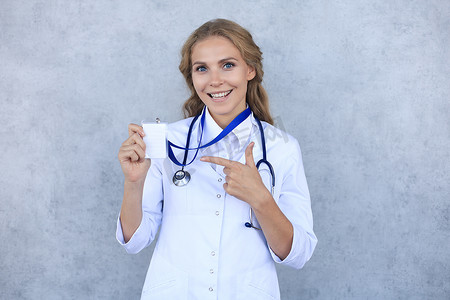 专业徽章摄影照片_微笑的金发女医生穿着制服站在灰色背景中，在徽章上显示她的名字。