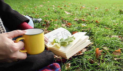 在温暖的一天，在绿色的草地上，一个女孩坐在秋天森林的毯子上，看书，喝着城市公园里黄色杯子里的热饮。