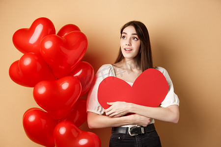 浪漫的女孩拥抱情人节大红心剪纸，站在浪漫气球附近，看着左边的标志，凝视着梦幻般的促销，米色背景