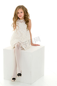 美丽的金发女孩穿着优雅的白色连衣裙坐在白色立方体上