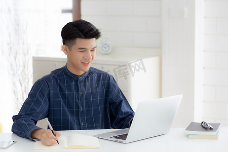 年轻的亚洲商人在笔记本上写作，规划工作，并在家里的办公桌上使用笔记本电脑，记录有关金融、男性学习、商业和沟通概念的笔记。