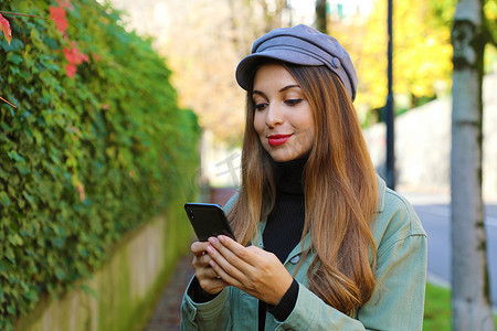 美丽的年轻女子，戴着帽子和夹克，在户外用手机发短信。