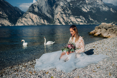 一个穿着漂亮白色连衣裙的女孩坐在加尔达湖的堤岸上。在意大利的山和湖的背景下拍摄了一个女人的照片。托尔博莱