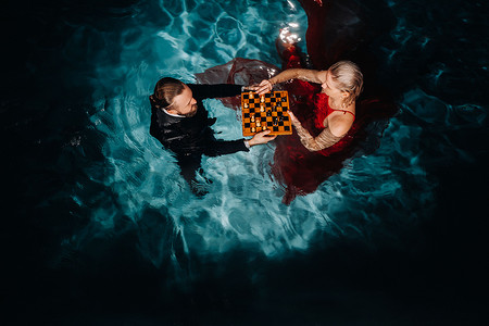 西装套装男摄影照片_一个穿西装的男人和一个穿红裙子的女孩在泳池的水上下棋