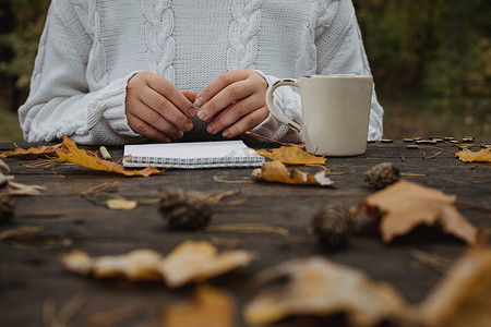 黄色页面摄影照片_身穿白色毛衣的年轻女子坐在公园一张黑色的旧桌子旁，端着一杯加咖啡的茶，在黄色秋叶的背景下读书写字。