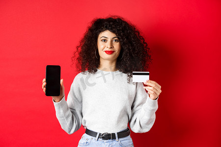 年轻的现代卷发女性展示塑料信用卡和手机屏幕，展示在线购物应用程序，站在红色背景上