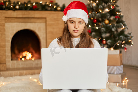 快乐的女人在圣诞树前看着笔记本电脑，戴着圣诞老人帽子的女士看着设备屏幕，通过视频通话祝贺某人，在客厅里摆出圣诞装饰品。