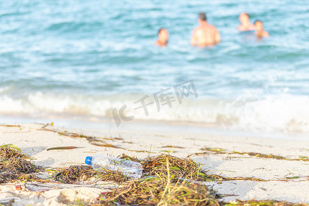 肮脏的海滩上空塑料水瓶的特写图像，肮脏的沙滩上装满了海藻、垃圾和废物，背景是海上的人们