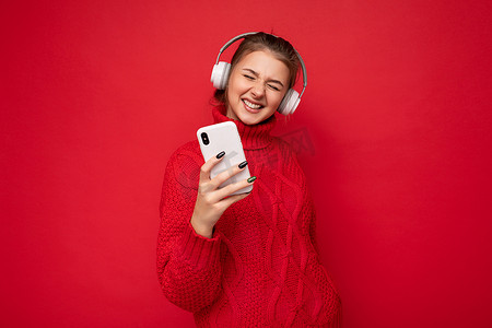 照片中，美丽快乐的黑发年轻女子穿着红色毛衣，与红色背景墙隔离，手持和使用手机，戴着蓝牙耳机玩得很开心