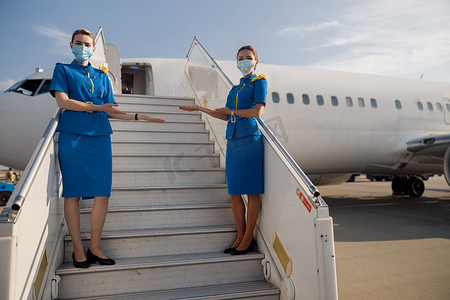 漂亮空姐摄影照片_两位身着蓝色制服、戴着防护面罩的漂亮空姐看着镜头、站在楼梯上欢迎乘客的全长镜头