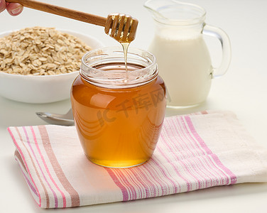 蜂蜜装在玻璃透明罐子里，木棍放在白桌上，在醒酒器后面，盘子里放着牛奶和燕麦片